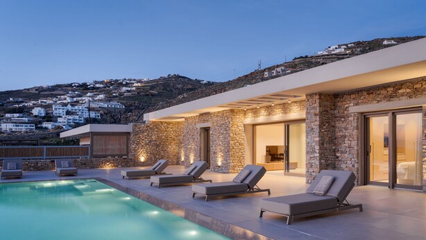 A modern poolside area with lounge chairs and a pool, overlooking a hillside with white buildings. - Beach House Houlakia Mykonos (Mykonos)