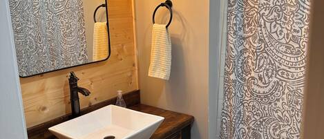 Un baño con tocador de madera, un lavabo blanco, un espejo con luces y un tocadiscos, un aro para toallas y una cortina de ducha estampada.