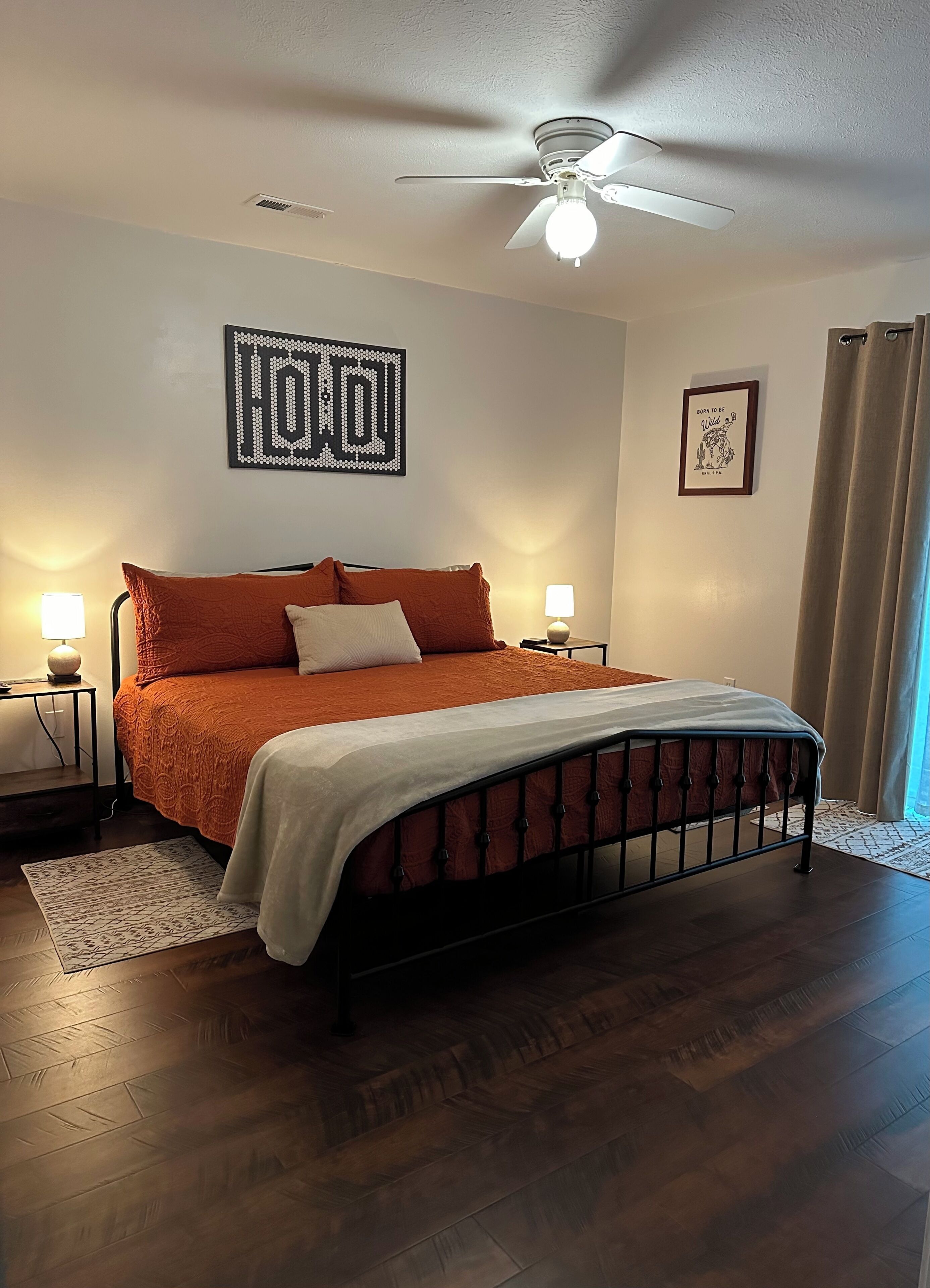 Un dormitorio con cama, ventilador de techo, una obra de arte en la pared, un cuadro enmarco y una ventana con cortinas.