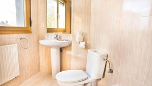 Une salle de bain comprenant des toilettes, un lavabo et une baignoire, avec des carreaux de couleur beige et une fenêtre.