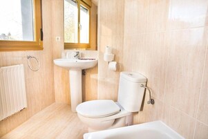 Un baño con inodoro, lavabo y bañera, con azulejos color beige y una ventana.