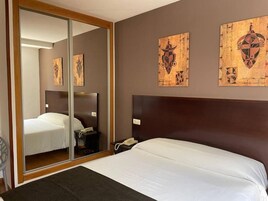 Habitación de hotel con cama, mesita de noche con teléfono, espejo y cuadro en la pared.