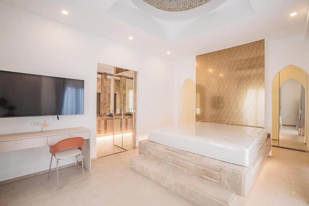 Een moderne slaapkamer met een groot bed, een bureau met stoel, een televisie en een badkamer zichtbaar door een glazen deur.