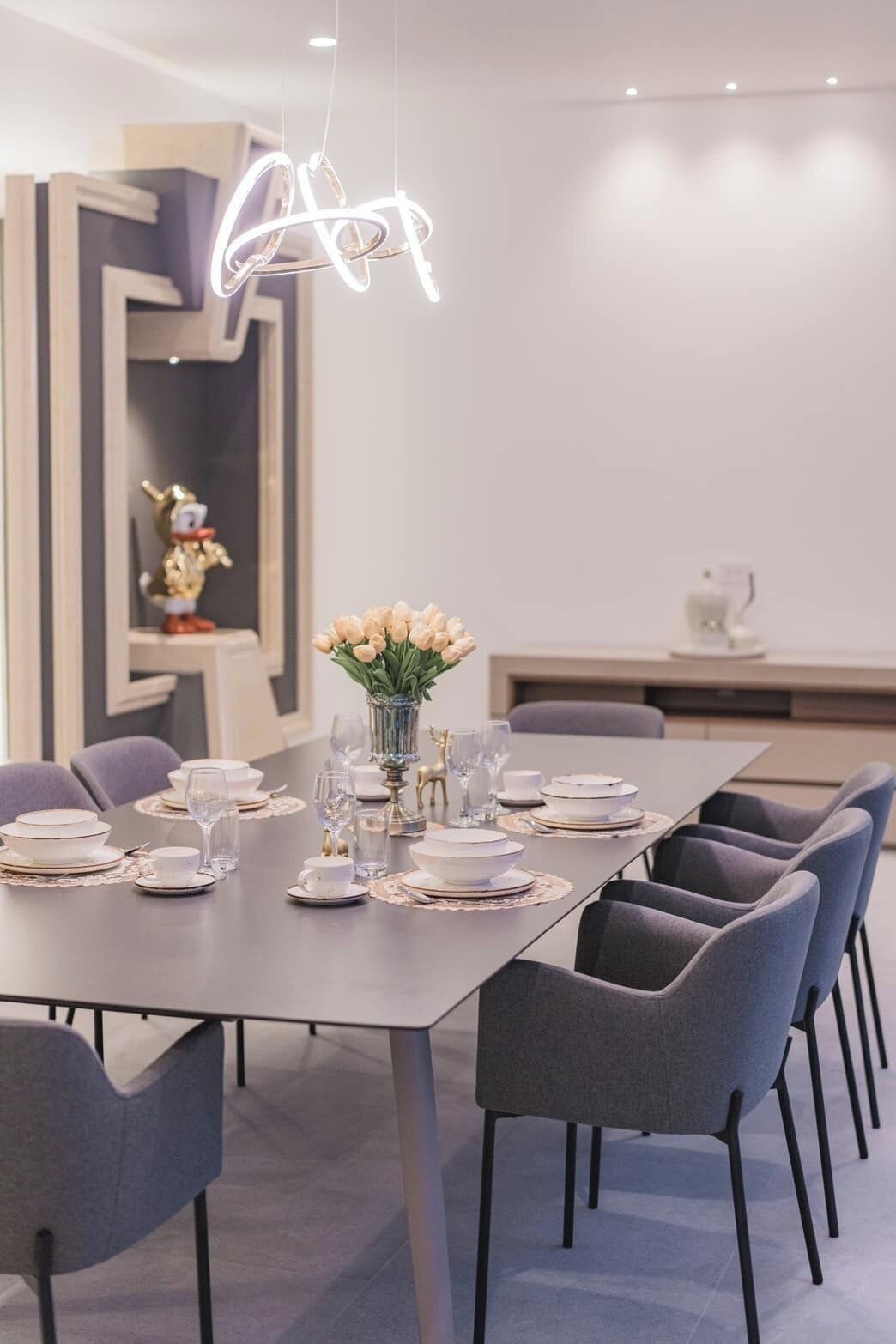 Un comedor moderno con una mesa larga dispuesta para una comida, sillas, un jarrón de flores y una lámpara de techo original.