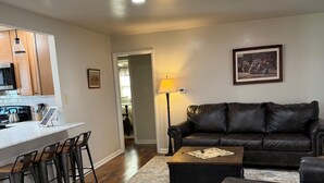 Ein Wohnzimmer mit einer schwarzen Ledersofa, einem Couchtisch und einem Teppich. Es gibt eine Küchenzeile mit Holzausbau und einer Mikrowelle.