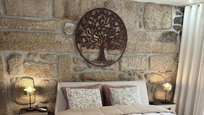 A bedroom with a stone wall, a decorative tree wall art, a bed with a beige quilt, two bedside tables with lamps, and a nightstand with drawers.