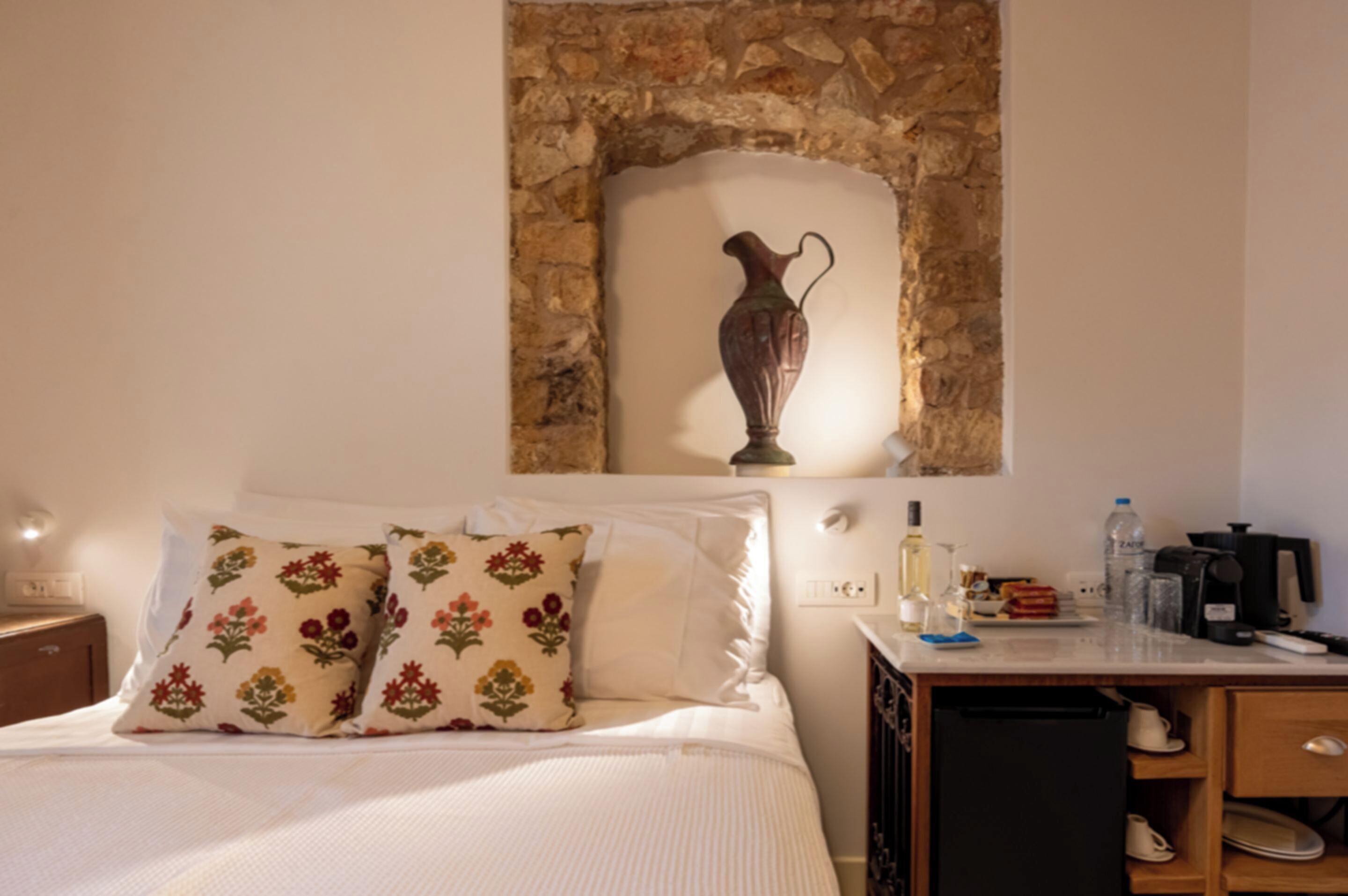 Un dormitorio con cama, almohadas, mesita de noche, cómoda, escritorio con lámpara y pared de piedra con un florero.