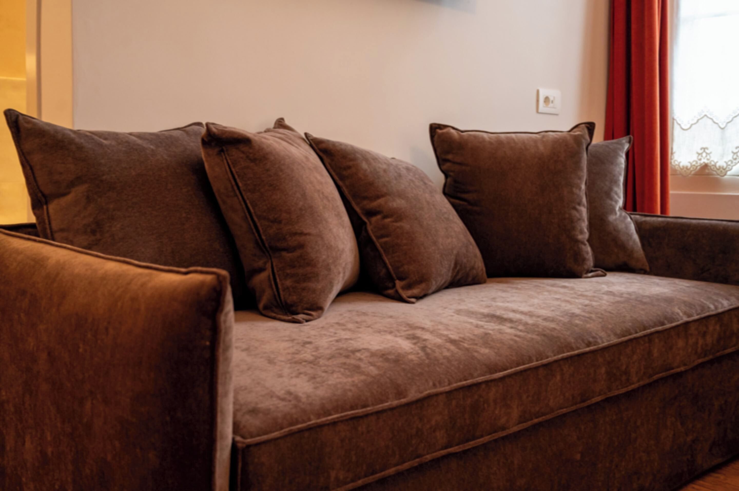 Un sofá marrón con varios cojines en una habitación con ventana y cortina roja.