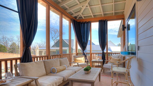 A covered outdoor seating area with a wooden deck, a sofa, chairs, a table, and a view of buildings in the background. - Luxury Pistache Park Retreat | Private Pool + Hot Tub | Carlton Landing (Carlton Landing)
