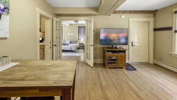 Una habitación con una mesa de comedor de madera, un televisor sobre un soporte, una cama y una puerta abierta que da a otra habitación.