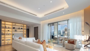 A modern living room with a sofa, a coffee table, and a dining area. There is a large window with a city view. - LUX  The Aurora Marina View Suite (Dubai)