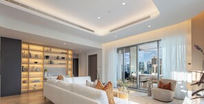 A modern living room with a sofa, a coffee table, and a dining area. There is a large window with a city view. - LUX  The Aurora Marina View Suite (Dubai)