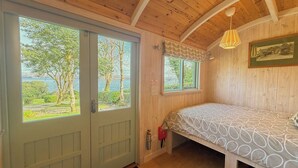A room with a bed, a window with a view of trees and water, a door with a view, and a ceiling with wooden beams.