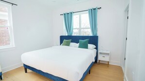 Ein modernes Schlafzimmer mit einem großen Bett, einem Nachttisch, einem Fenster mit Vorhängen und einer sichtbaren Ziegelwand.