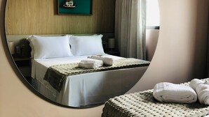 Quarto de hotel com um espelho grande refletindo a cama, uma mesinha com abajur, um quadro na parede e um aparelho de ar condicionado.