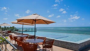 Área de piscina ao ar livre com espreguiçadeiras e guarda-sóis, com vista para um oceano azul e límpido.