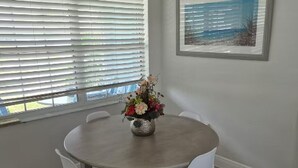 A dining area with a round table, white chairs, a vase of flowers, a window with blinds, and a framed picture on the wall. - Baby Blue! (Port Charlotte)