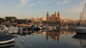 Una marina con barcos amarrados, una iglesia grande con dos cúpulas y una superficie de agua tranquila que refleja las construcciones.