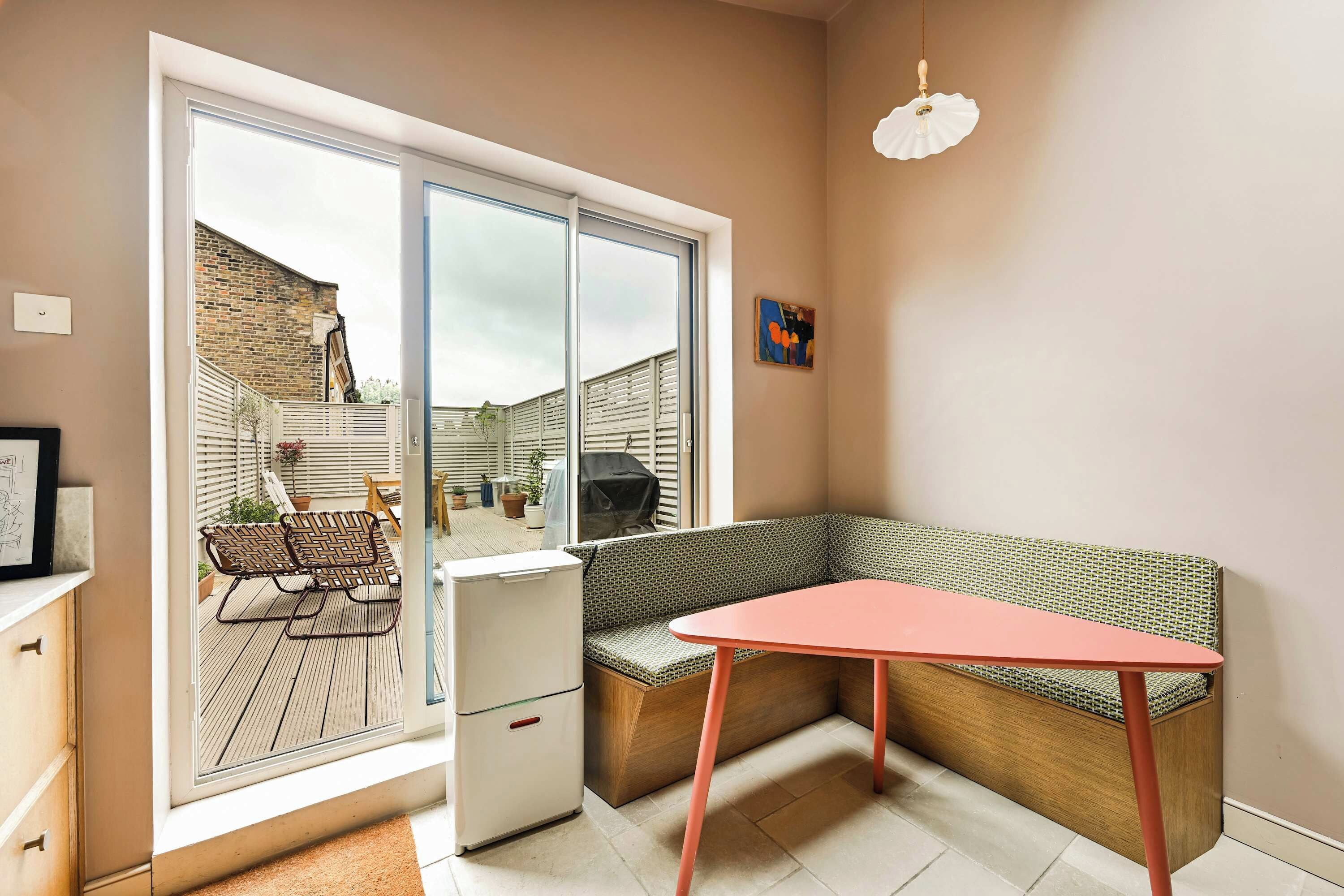 Une chambre moderne dotée d’une grande porte-fenêtre coulissante, d’un banc en bois avec un siège rembourré, d’un petit réfrigérateur blanc et d’une table rouge.
