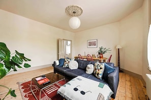 A living room with a sofa, a coffee table, a red rug, a plant, and a dining area with chairs and a table. - The Sun Deck Townhouse (Greater London)