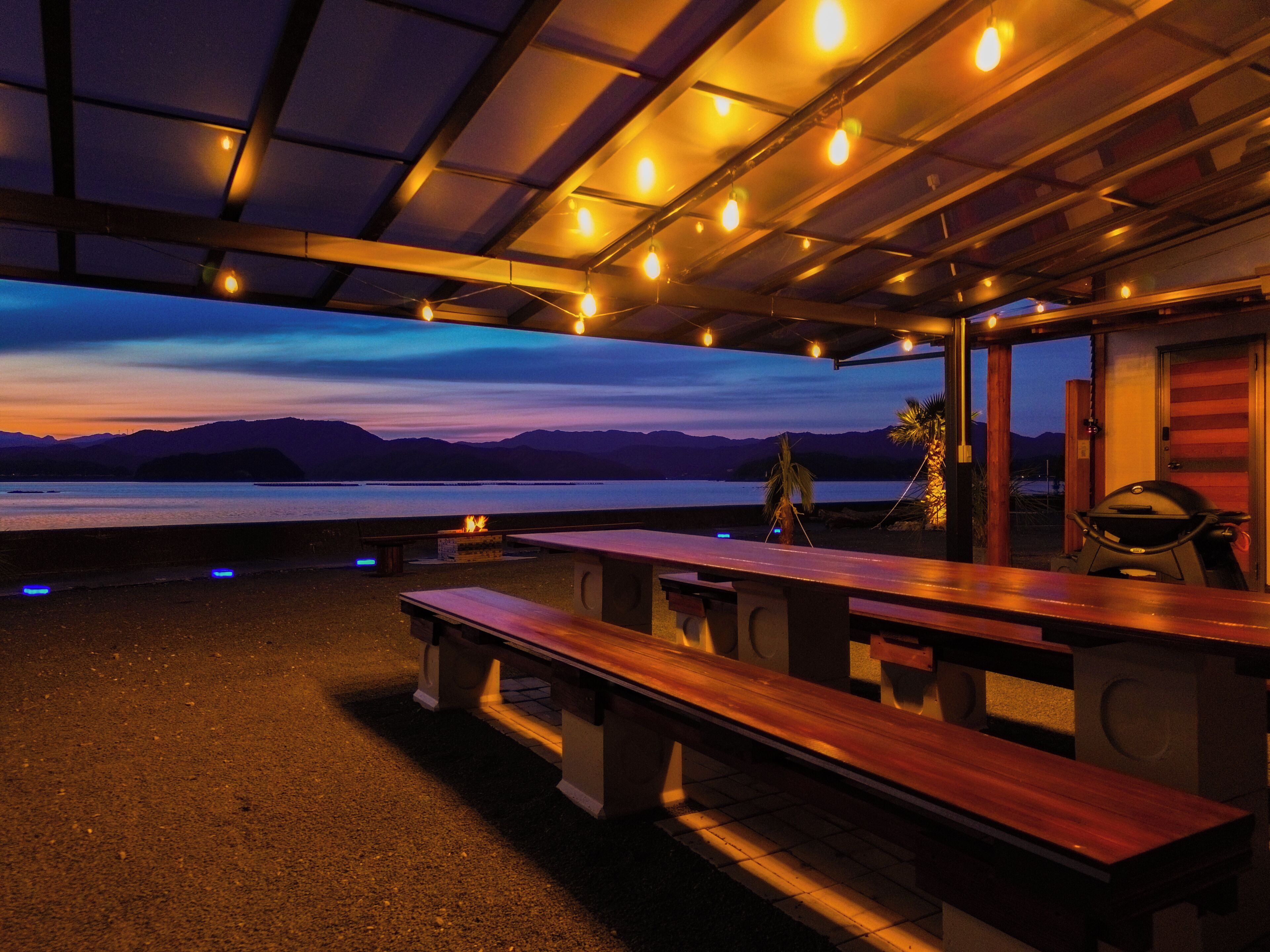 An outdoor seating area with wooden benches and tables, a grill, and a view of a body of water with mountains in the background.