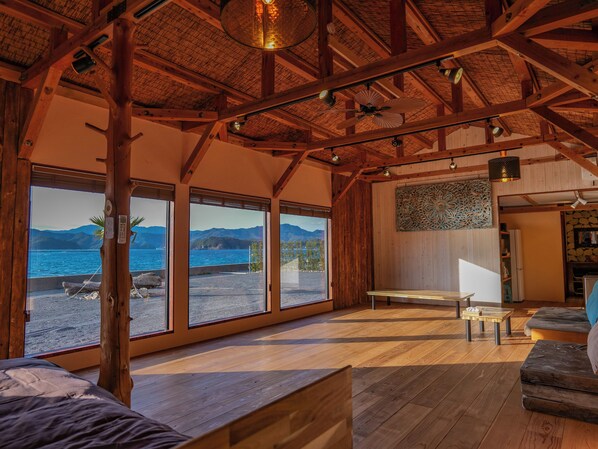 A wooden interior with large windows, a ceiling fan, and a view of a beach.