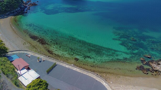 An aerial view of a coastal area with clear waters, a beach, and a small building with a red roof.