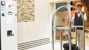 A hotel lobby with a self-service kiosk, a luggage cart, and a person standing by a door.