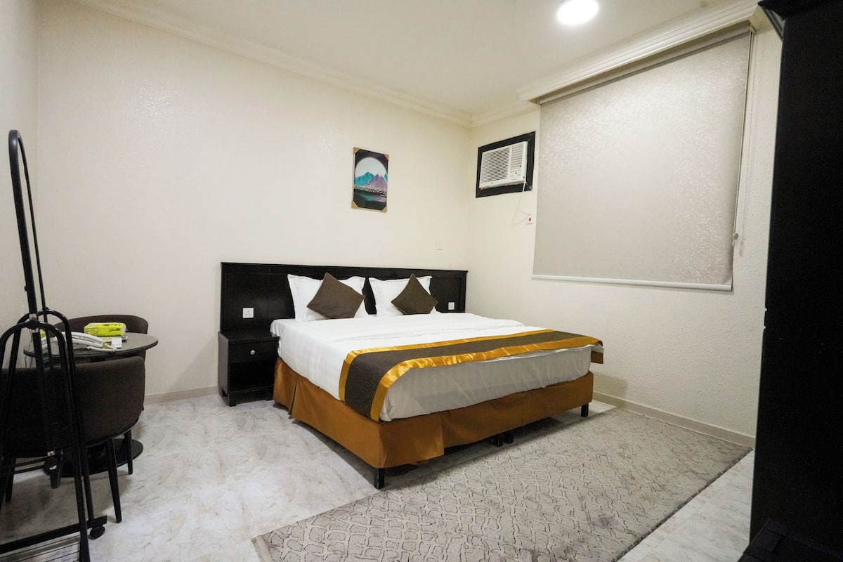 Un dormitorio con cama, escritorio, silla, un cuadro en la pared y ventana.