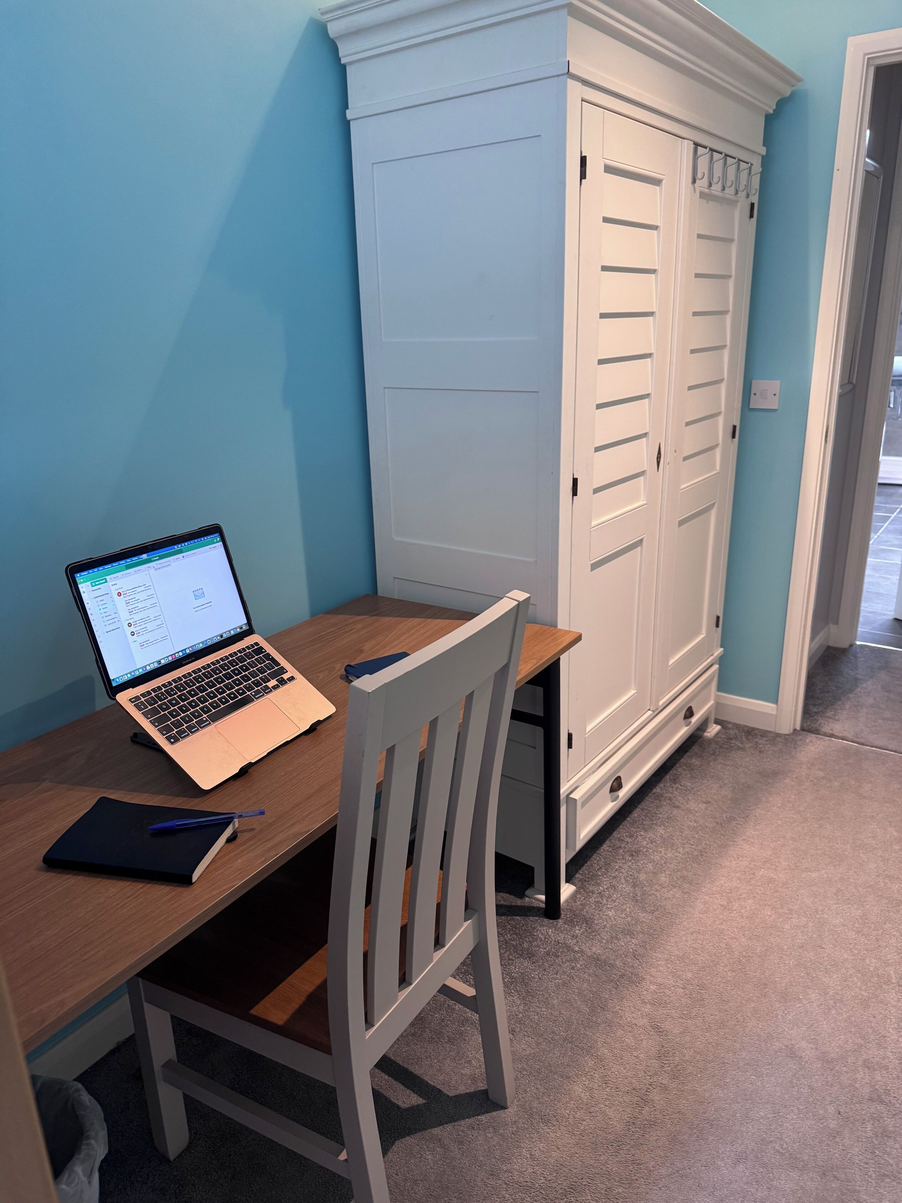 A laptop on a wooden desk with a chair, a notebook, and a pen. There is a white wardrobe with multiple doors and a light blue wall in the background.