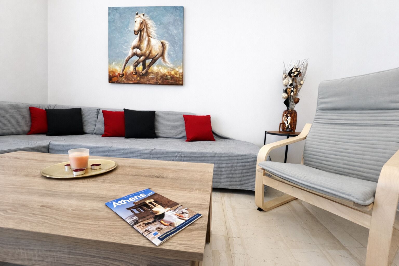 A modern living room with a grey sofa, red and black cushions, a wooden coffee table with a magazine, a glass of drink, and a painting of a horse.