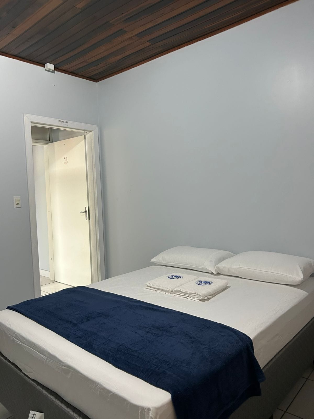 A bedroom with a bed, a blue blanket, white pillows, and a white door with a number.