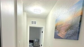Een gang met een deur naar een kamer, een plafondlamp en een schilderij aan de muur.