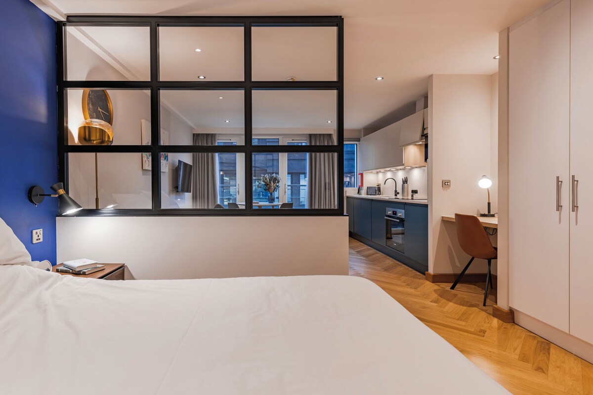 A modern bedroom with a large bed, a desk, and a view of the city through a window.