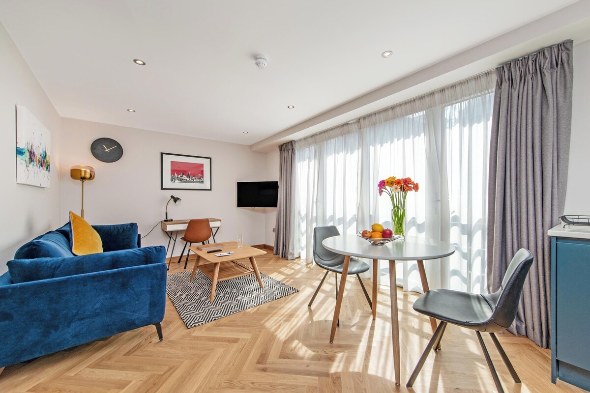 A modern living room with a blue sofa, a dining table with chairs, a TV, and a large window with curtains.