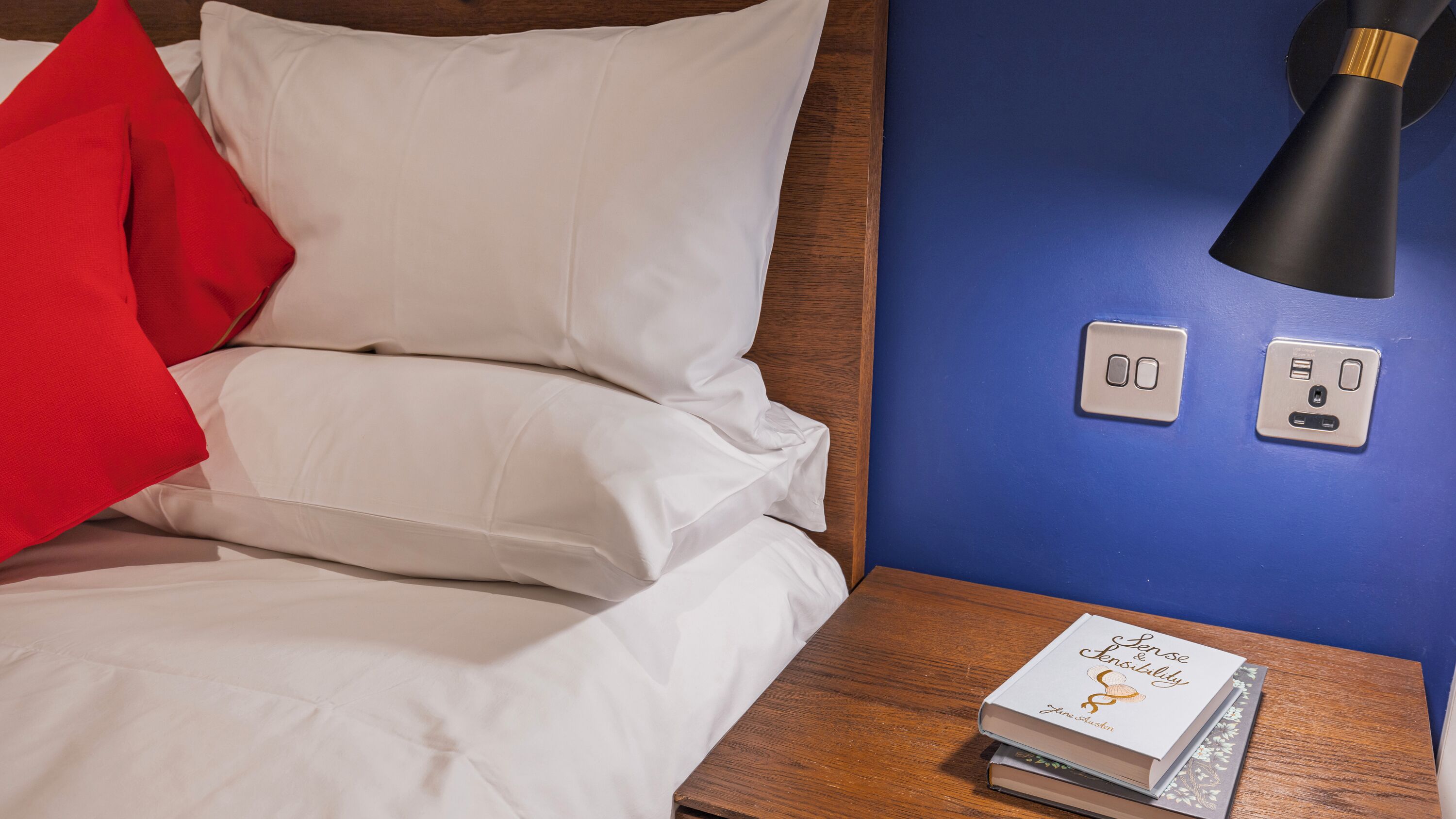 Een net opgemaakt bed met witte lakens en een rode kussen, een nachtkastje met een boek getiteld 'Sense & Sensibility', en een wandlamp.