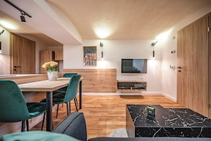 A modern living room with a wooden floor, a dining area with green chairs, a TV, and a wooden wardrobe.