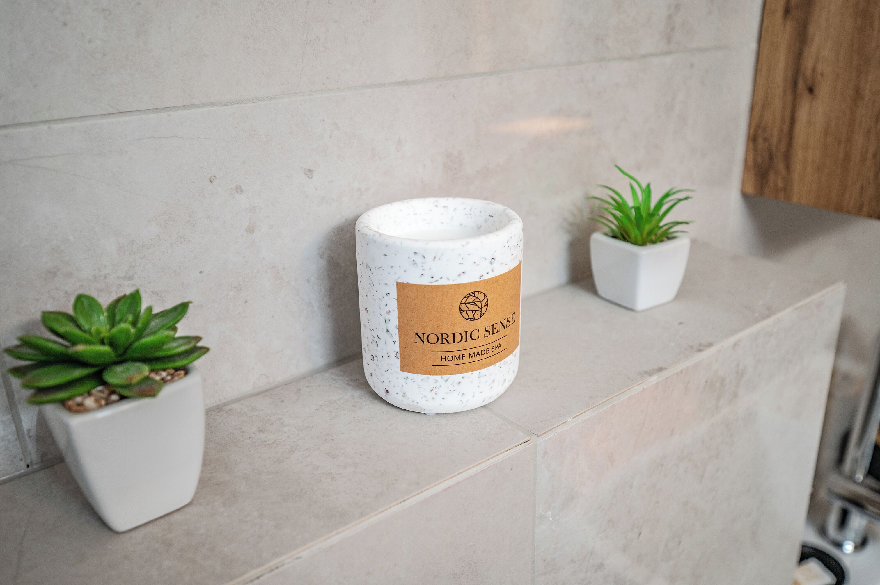 A spa candle labeled 'Nordic Sense' on a tiled surface with two potted plants.