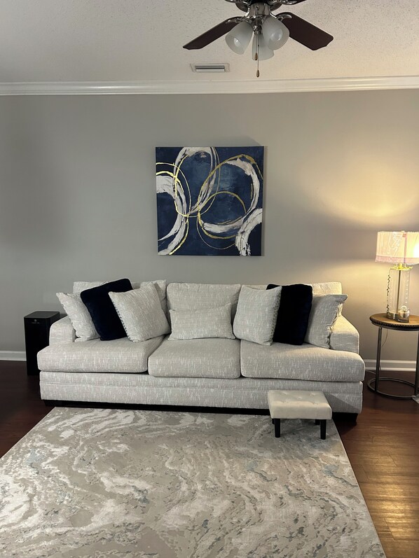 Un salon avec un canapé gris, un tapis en marbre, un ventilateur de plafond et un tableau abstrait accroché au mur.