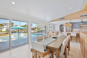A modern kitchen with wooden cabinetry and a large island, a dining area with a wooden table and chairs, and a view of a pool and resort buildings. - Pink Pearl Waterfront Retreat | Canal + Dock (Holmes Beach)