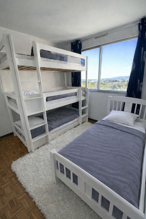 Une chambre avec des lits superposés, un lit simple, une vue par la fenêtre et une moquette blanche.