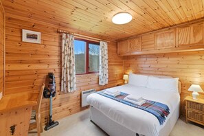 A wooden-paneled room with a bed, a window with curtains, a heater, and a lamp on the nightstand.