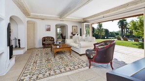 A spacious living room with a large window, a white sofa, a patterned rug, and a red armchair. - Lovely family villa in Saint Paul de Vence (La Colle-sur-Loup)