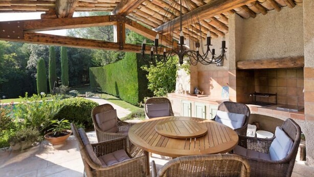 An outdoor patio with wicker furniture, a round table, a stone fireplace, and a view of a garden. - Lovely family villa in Saint Paul de Vence (La Colle-sur-Loup)