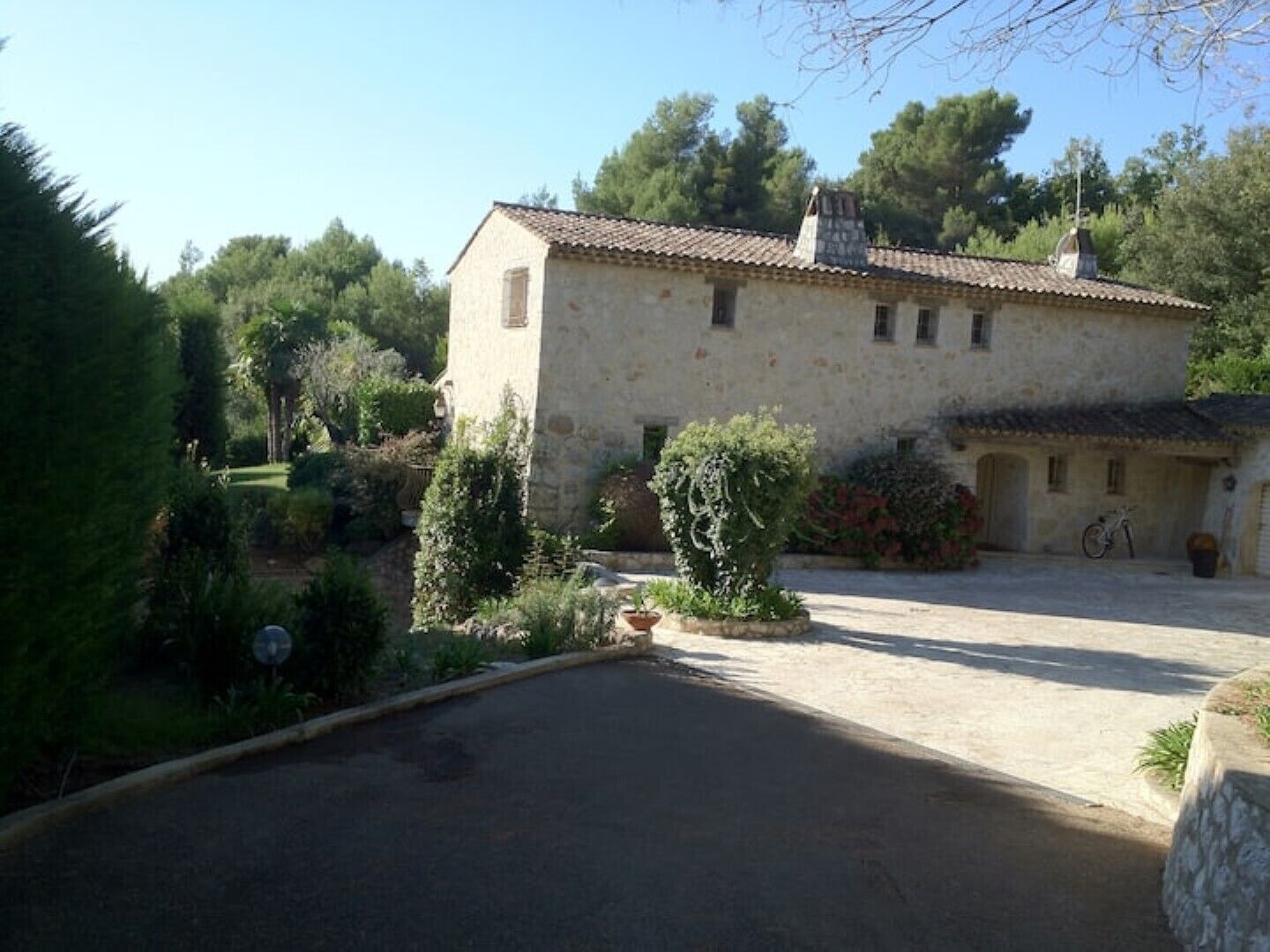 A stone building with a tiled roof, surrounded by greenery and a paved driveway.