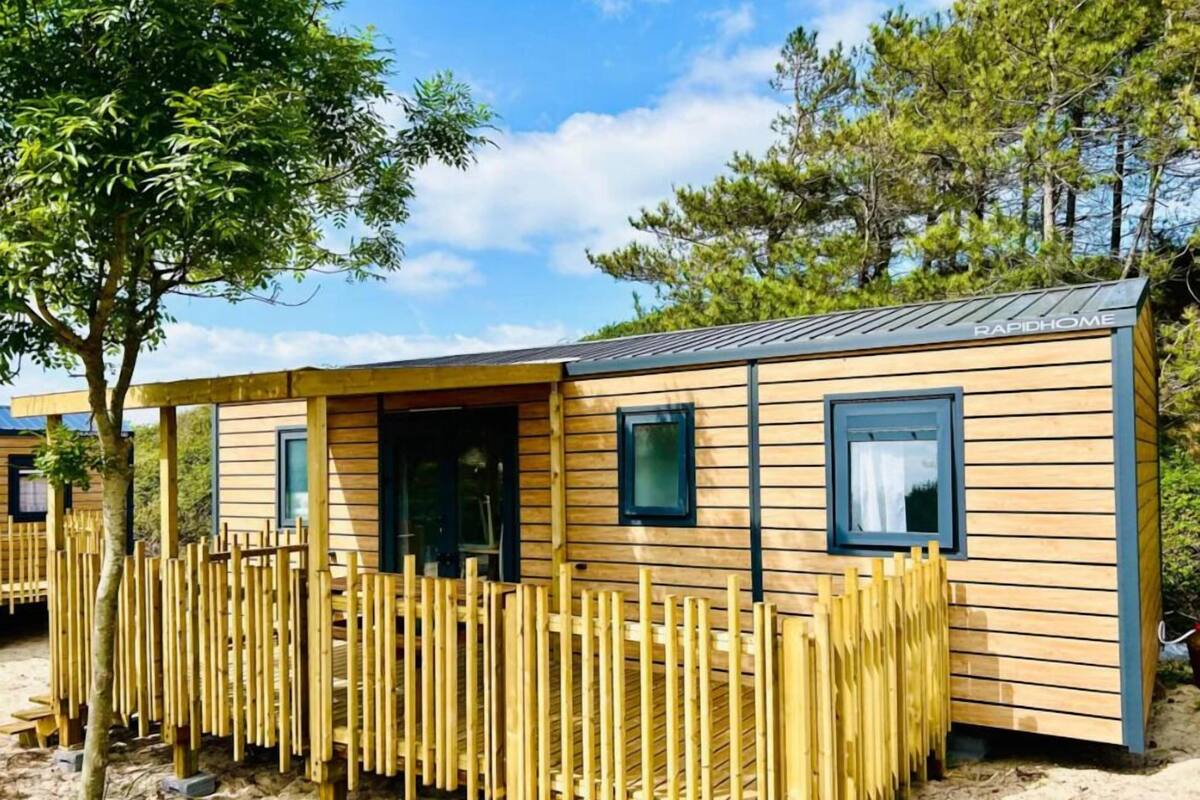 A wooden mobile home with a fenced outdoor area, windows, and a clear sky.