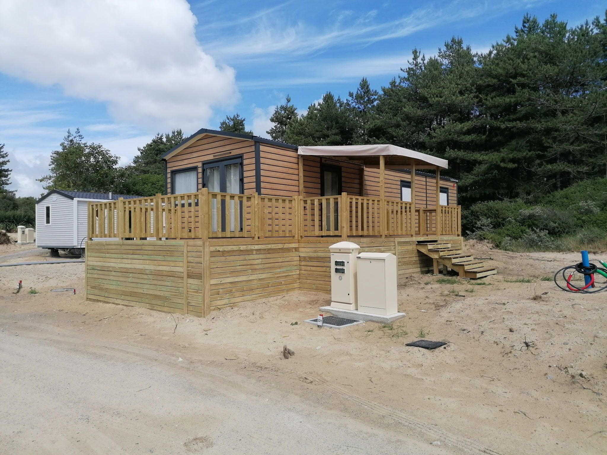 Une cabane en bois avec une terrasse entourée d’une clôture, un petit abri attenant et un coffret de raccordement.