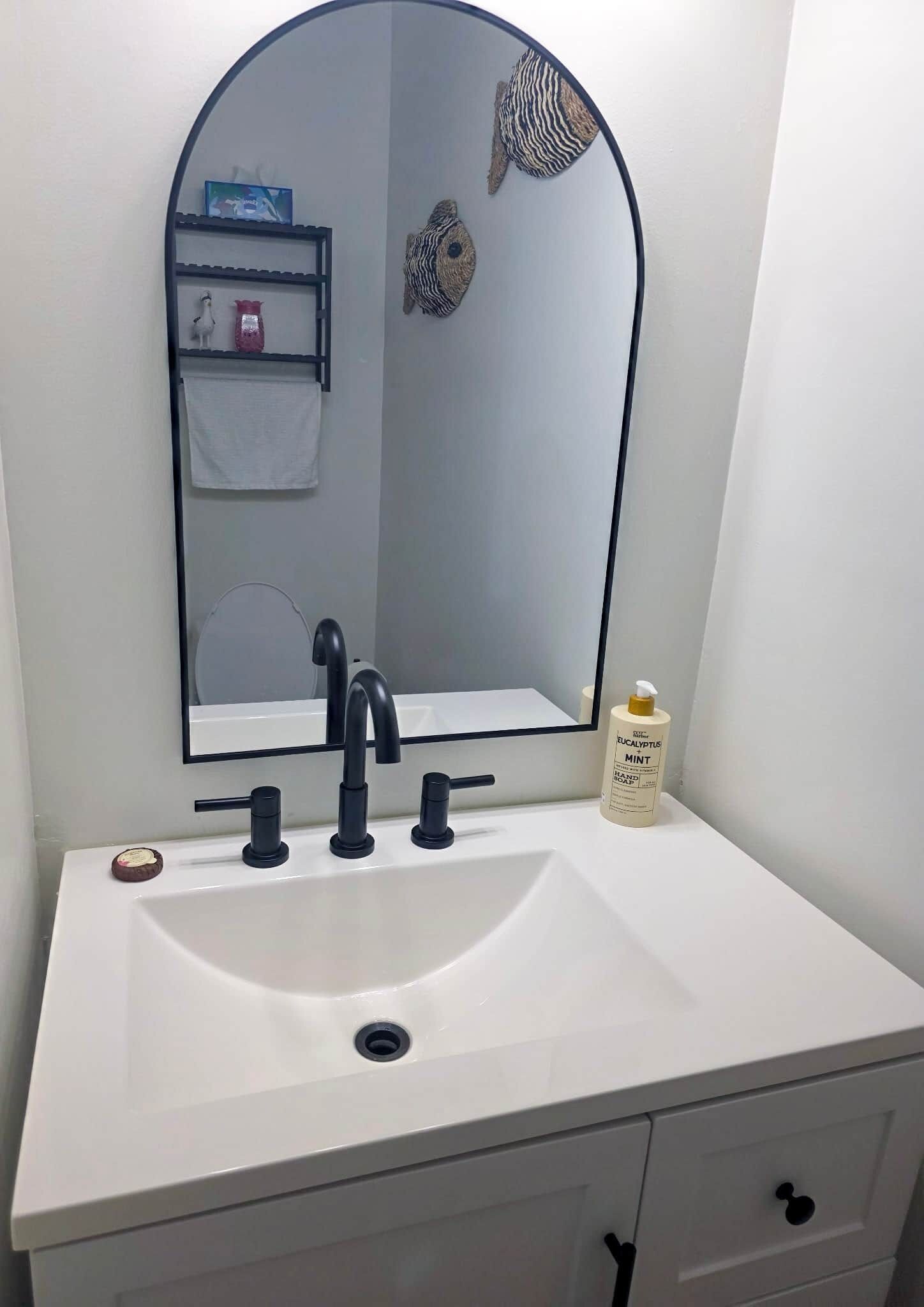 A bathroom with a white sink, black faucet, and a mirror above it. There is a shelf with a towel and a bottle of hand soap.