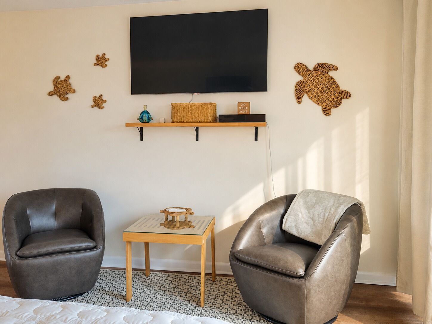 A living room with two leather armchairs, a small wooden table, a TV mounted on the wall, and a shelf with decorative items.