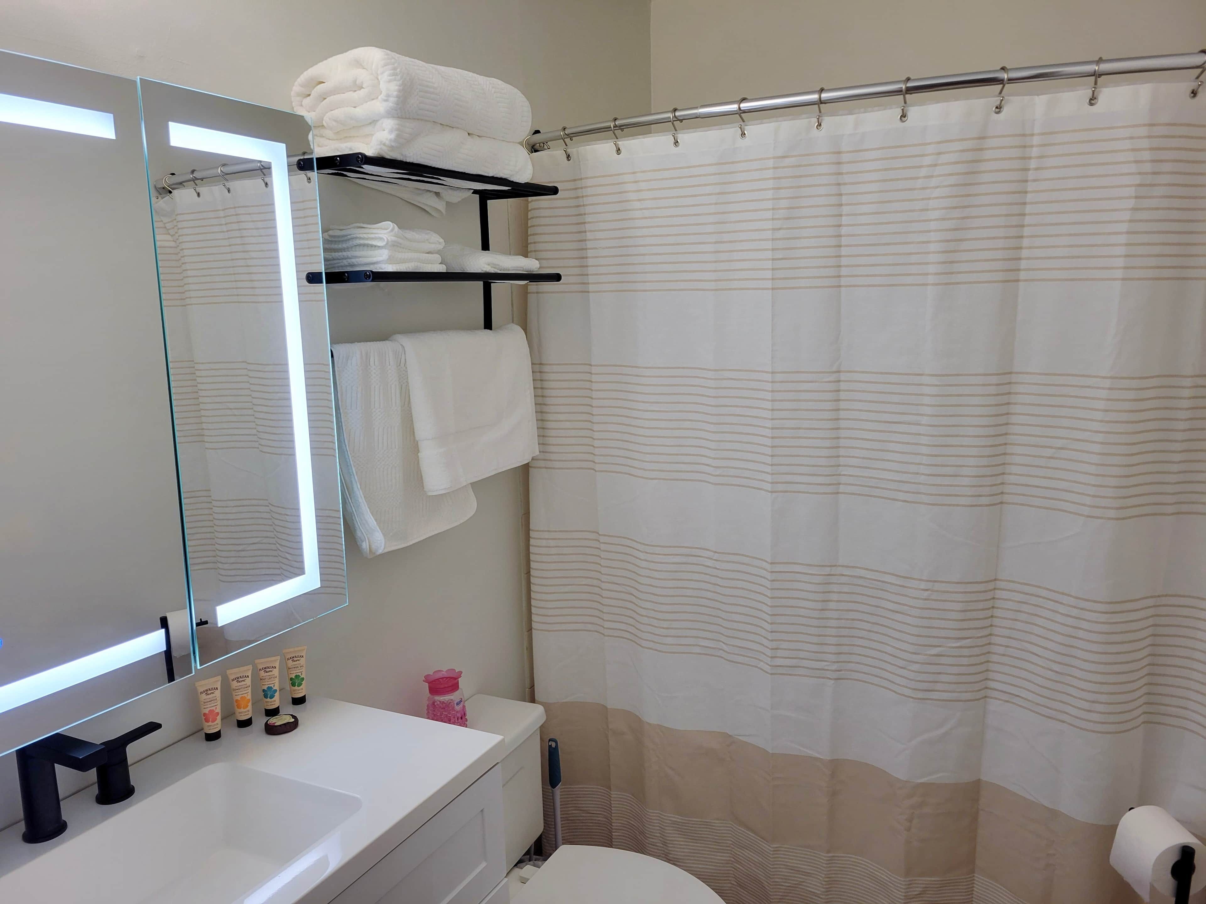 Een badkamer met een wit wastafelmeubel, een spiegel, een handdoekrek met opgevouwen handdoeken, een douchegordijn en toiletartikelen op het aanrecht.